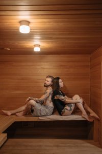 A man and woman sit back-to-back inside a wooden sauna.